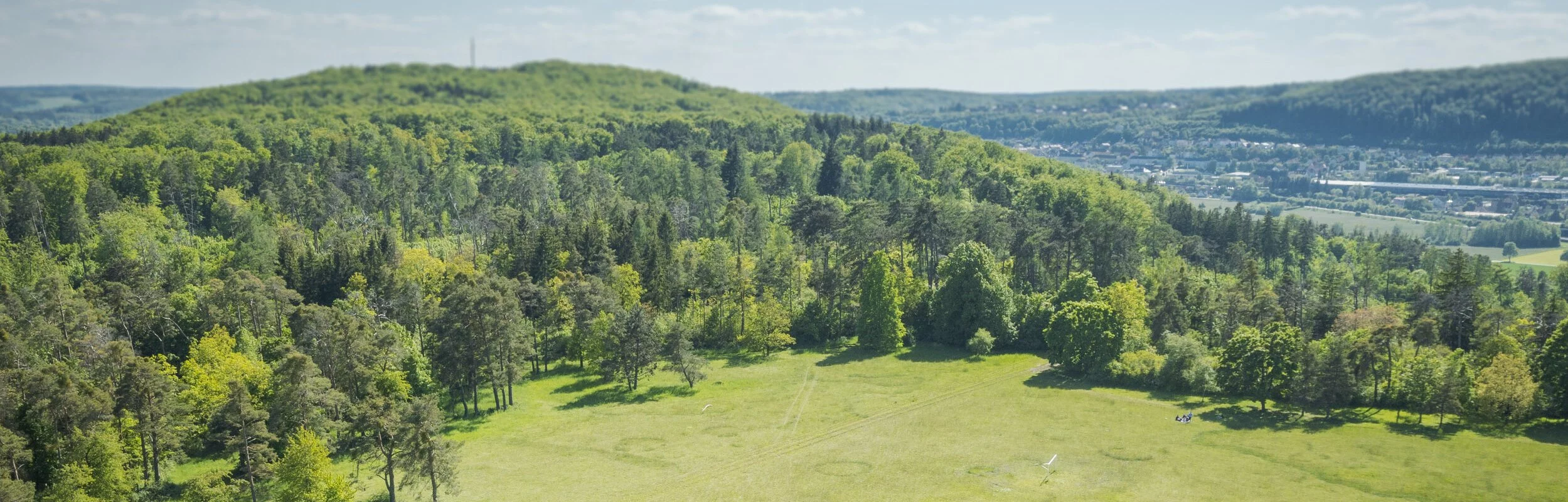 Luftaufnahme eines kleinen Hauses auf einer Wiese. Großer Weitblick mit viel Wald und Grün.